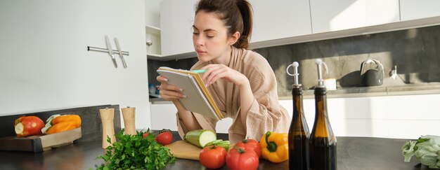 Retrato de una mujer revisando la lista de compras mirando verduras sosteniendo una receta de lectura de cuaderno mientras