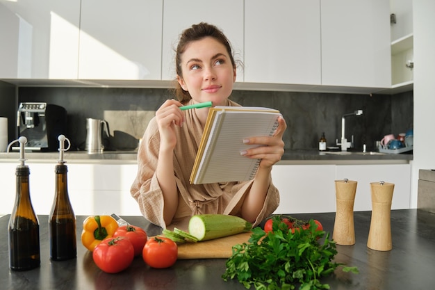 Foto gratuita retrato de una mujer revisando notas de recetas en un cuaderno de pie en la cocina con verduras cocinando comida