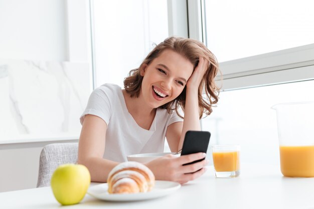 Retrato de mujer riendo en camiseta blanca revisando noticias en el teléfono móvil mientras está sentado en la mesa de la cocina