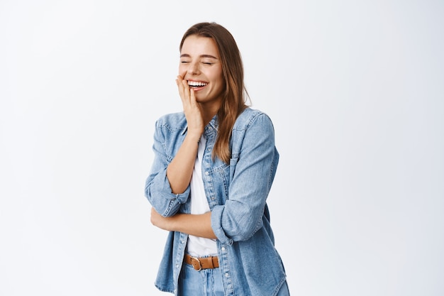 Retrato de mujer riendo sintiéndose feliz, divirtiéndose, cerrando los ojos y riendo de una broma divertida, de pie en ropa casual contra la pared blanca