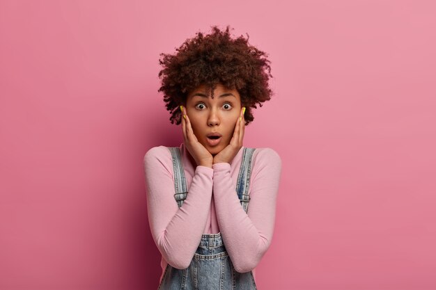 Retrato de mujer rizada asombrada agarra la cara y mira con ojos saltones, chismes sobre algo increíble, vestida de manera informal, posa contra la pared rosa, está ansiosa por un terrible accidente.