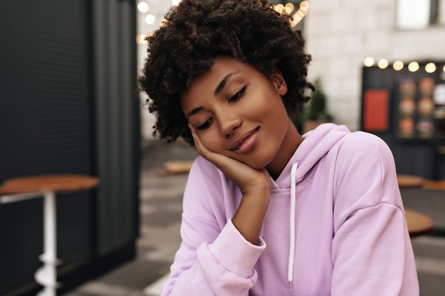 Retrato de mujer rizada morena encantadora tranquila en sudadera con capucha púrpura sonriendo suavemente con los ojos cerrados afuera