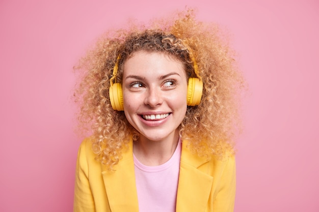 Retrato de mujer rizada sonriente feliz disfruta de la lista de reproducción favorita escucha música a través de auriculares inalámbricos mira hacia otro lado sonríe a la cámara