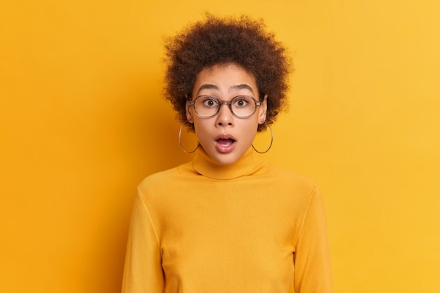 Retrato de mujer rizada sorprendida mira con la boca abierta reacciona ante noticias asombrosas usa gafas transparentes pendientes de cuello alto. Adolescente afroamericano impresionado sin palabras mantiene la boca abierta