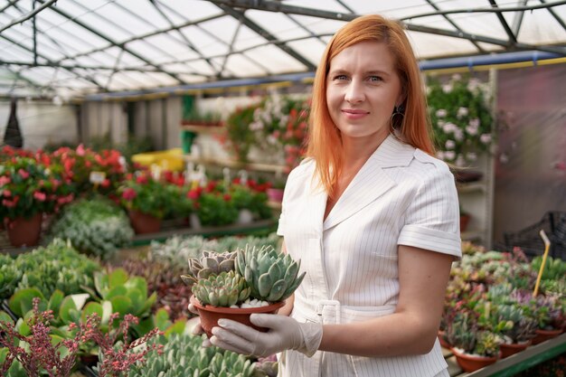 Retrato de mujer rojiza con guantes de goma y ropa blanca sosteniendo suculentas o cactus en macetas con otras plantas verdes