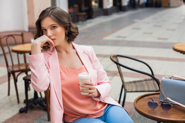 Retrato de mujer romántica con estilo sentada en la cafetería tomando café, vestida con chaqueta y blusa rosa, tendencias de color en la ropa, moda primavera verano, accesorios, gafas de sol y bolso, pensativo