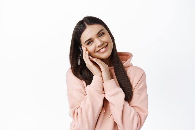 Retrato de mujer romántica sonriente apoya su mejilla en las manos y mirando coqueta al frente, sonrojándose de felicidad, de pie contra la pared blanca