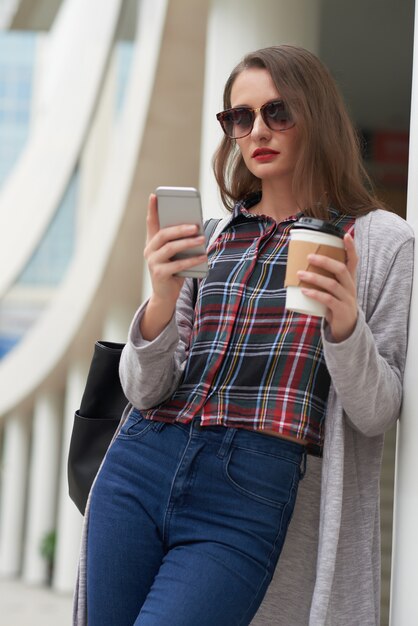 Retrato de mujer en ropa casual usando el teléfono inteligente mientras bebe café al aire libre