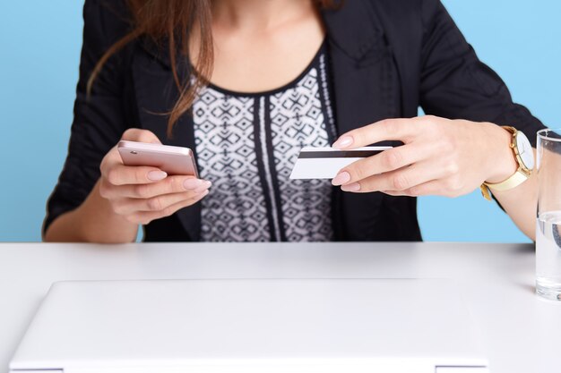 Retrato de mujer sin rostro viste traje negro con teléfono celular y tarjeta de crédito mientras está sentado en la mesa blanca cerca de la computadora portátil y un vaso de agua, aislado sobre la pared azul. Concepto de pago en línea