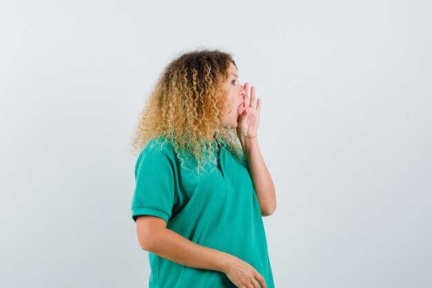 Retrato de mujer rubia con cabello rizado diciendo secreto detrás de la mano en camiseta verde y mirando emocionado vista frontal