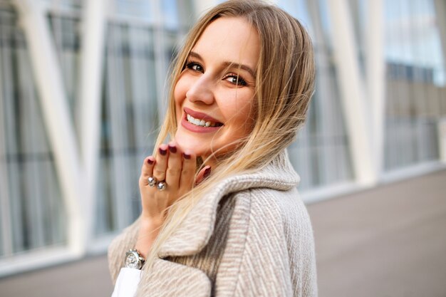 Retrato de mujer rubia elegante posando en la calle con abrigo, maquillaje natural, rostro sensual y largos cabellos rubios, clima otoñal ventoso de cerca.