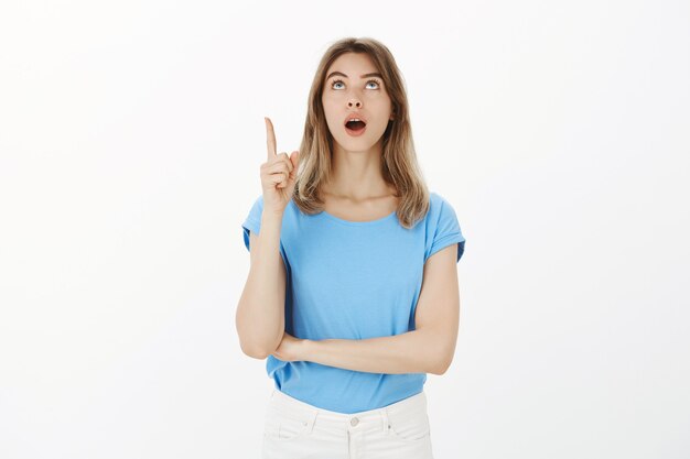 Retrato de mujer rubia emocionada y asombrada dejar caer la mandíbula, señalar con el dedo y mirar hacia arriba fascinado