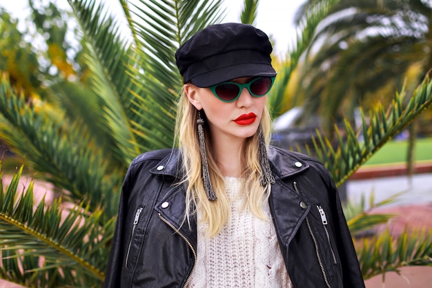 Retrato de mujer rubia con estilo posando cerca de palmeras en invierno de cerca, con chaqueta de cuero, gafas de sol de moda retro, gorra y pendientes largos, colores suaves.