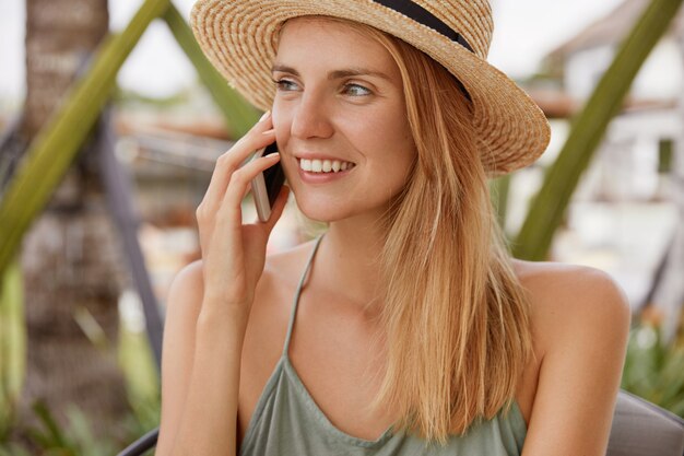 Retrato de mujer rubia feliz usa sombrero de paja y sonrisa feliz, mira positivamente hacia otro lado, tiene una conversación agradable con su novio a través del teléfono celular, comparte impresiones sobre estar en un resort de playa. La gente y hablar