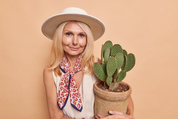 Retrato de mujer rubia guapa con maquillaje mínimo, comlexión bien cuidada sostiene olla de cactus viste sombrero vestido pañuelo atado aislado sobre pared beige