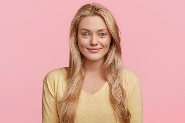 Retrato de mujer rubia hermosa alegre con mirada complacida, viste suéter casual amarillo, modelos contra la pared rosa. La hembra joven contenta tiene una sonrisa agradable en la cara. Tiro horizontal