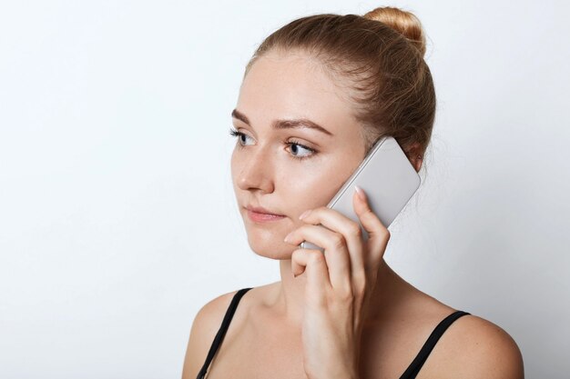 Retrato de mujer rubia mirando a un lado con expresión seria mientras llama a su amigo o familiares, escuchando atentamente Joven mujer atractiva chateando por teléfono móvil.