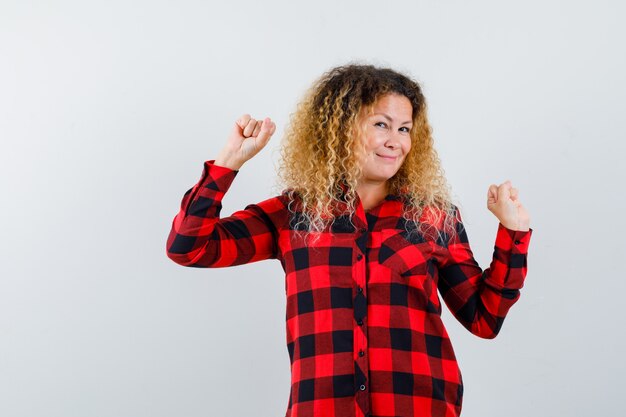 Retrato de mujer rubia con pelo rizado mostrando gesto ganador en camisa a cuadros y mirando divertido vista frontal