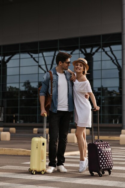 Retrato de mujer rubia con sombrero y hombre moreno con chaqueta de mezclilla pantalones negros sonriendo sinceramente Los turistas posan con maletas cerca del aeropuerto en el cruce de peatones