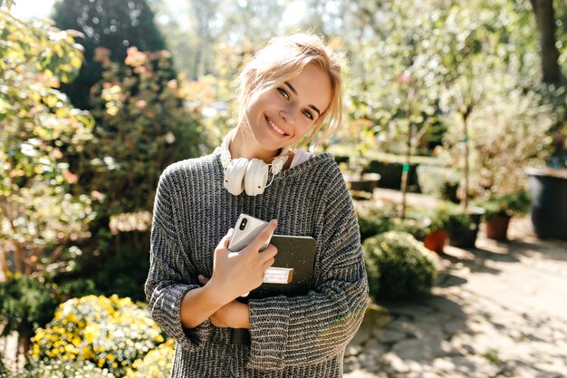 Retrato de mujer rubia sonriente en suéter de punto con teléfono y portátil en sus manos.