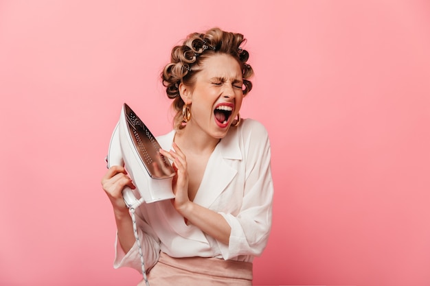 Foto gratuita retrato de mujer con rulos, tocando el hierro y gritando de dolor en la pared rosa