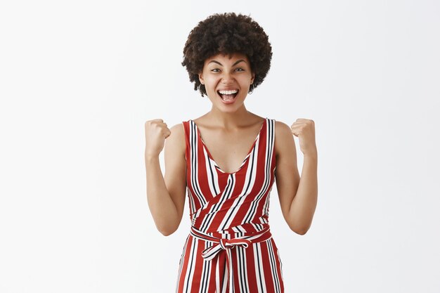 Retrato de mujer satisfecha y guapa triunfante con corte de pelo afro taising puños cerrados en gesto de celebración sintiéndose feliz por ganar o victoria sonríe ampliamente sobre la pared gris