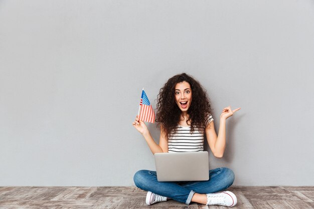 Retrato de mujer satisfecha con hermosa sonrisa sentada en postura de loto con computadora plateada en las piernas sosteniendo la bandera americana en la mano sobre la pared gris