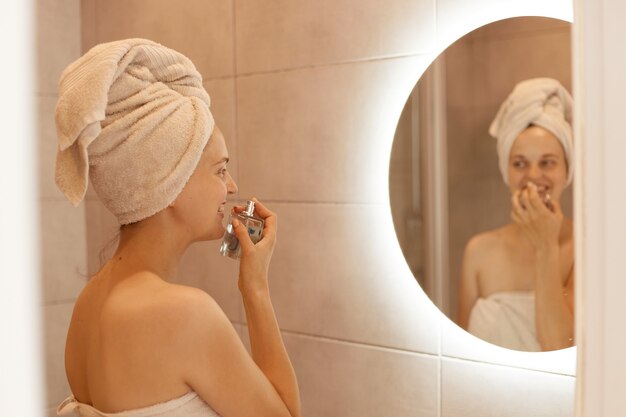 Retrato de mujer satisfecha oliendo perfume mientras posa en el baño, disfrutando de un olor agradable, de pie con los hombros desnudos y una toalla blanca sobre su cabello frente al espejo.