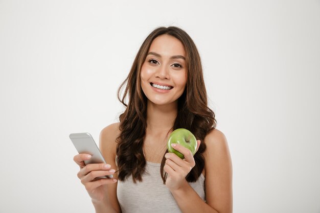 Retrato de mujer satisfecha con una sonrisa perfecta usando un teléfono inteligente plateado y comiendo manzana verde fresca aislada sobre la pared blanca