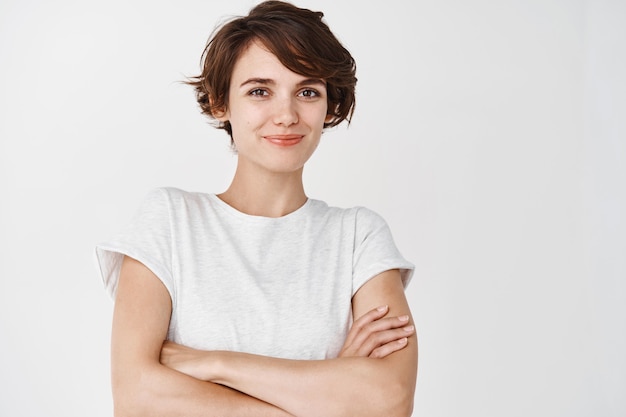 Retrato de mujer segura y feliz con pelo corto, brazos cruzados sobre el pecho como profesional y sonriente, de pie contra la pared blanca