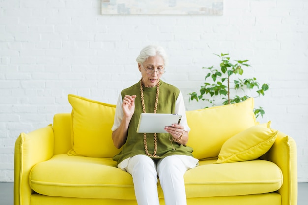 Retrato de mujer senior sentado en el sofá amarillo mirando tableta digital