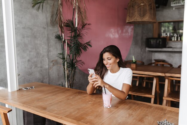 Retrato de mujer sentada en la cafetería y charlando con una sonrisa en el smartphone