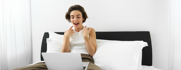 Foto gratuita retrato de una mujer sentada en la cama con una computadora portátil jadeando y mirando sorprendida a la pantalla de la computadora