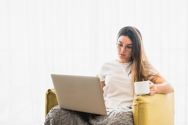 Retrato de mujer sentada en un sillón sosteniendo una taza de café usando una computadora portátil