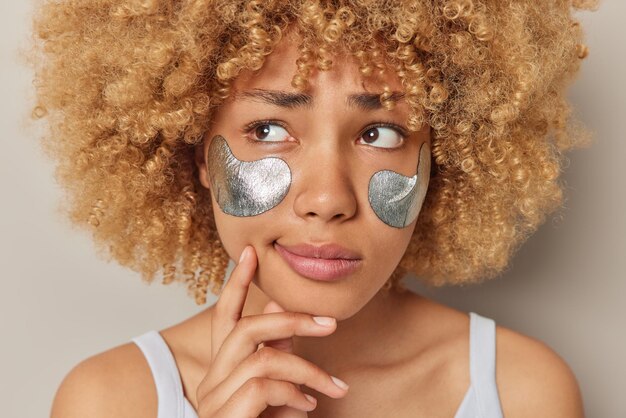 El retrato de una mujer seria y pensativa con el pelo rizado y tupido aplica parches de belleza plateados debajo de los ojos para eliminar la hinchazón y las arrugas y los hombros desnudos contra el fondo gris del estudio