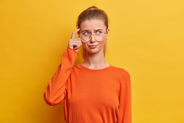 Retrato de mujer seria y reflexiva mantiene el dedo en la sien, intenta concentrarse en algo, viste un jersey naranja casual, posa