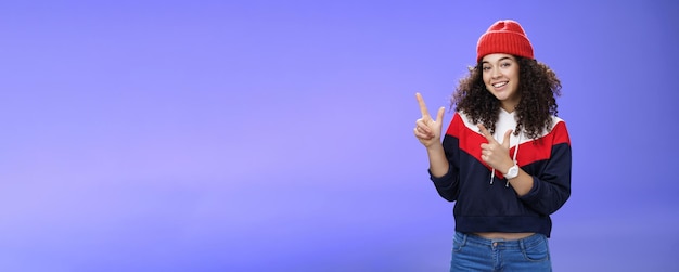 Foto gratuita retrato de una mujer simpática y guapa con el pelo rizado en un gorro rojo cálido apuntando a la esquina superior izquierda