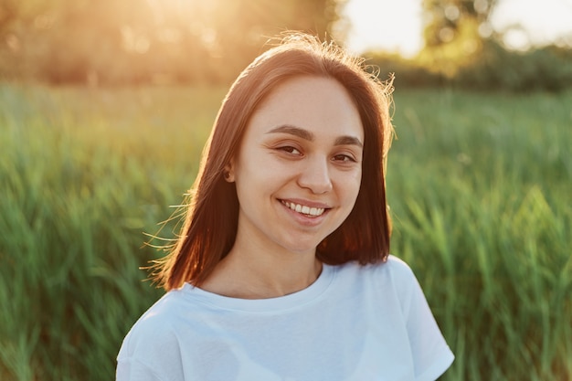 Retrato de mujer sonriente adulta joven con ropa blanca mirando directamente a la cámara con expresión feliz, posando en el prado verde al atardecer o al amanecer.