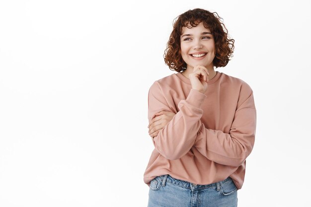 Retrato de mujer sonriente atractiva y segura, sostenga la mano en la barbilla, mirando la esquina superior izquierda con cara determinada y satisfecha, mirando algo bueno, asiente con aprobación, fondo blanco.