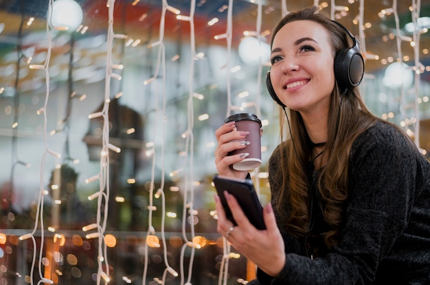 Retrato de mujer sonriente con auriculares con taza y teléfono cerca de luces de Navidad