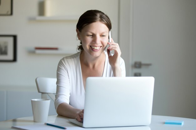 Retrato de mujer sonriente cerca de portátil hablando por teléfono.