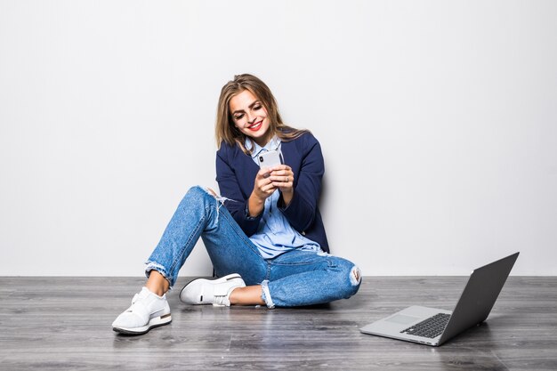 Retrato de mujer sonriente escribiendo mensajes de texto o desplazándose en internet mediante teléfono móvil, sentado sobre una pared gris