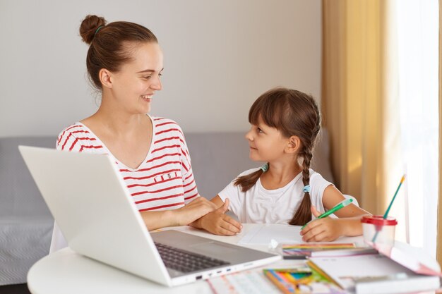 Retrato de mujer sonriente feliz vistiendo ropa casual ayudando a su hija con lecciones, mujer mirando a su hijo con amor, sentada a la mesa con libros y portátil.