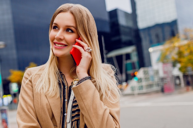 Retrato de mujer sonriente hablando por teléfono móvil de cerca. Señora de negocios exitosa con smartphone. Accesorios con estilo. Abrigo beige. Edificios urbanos