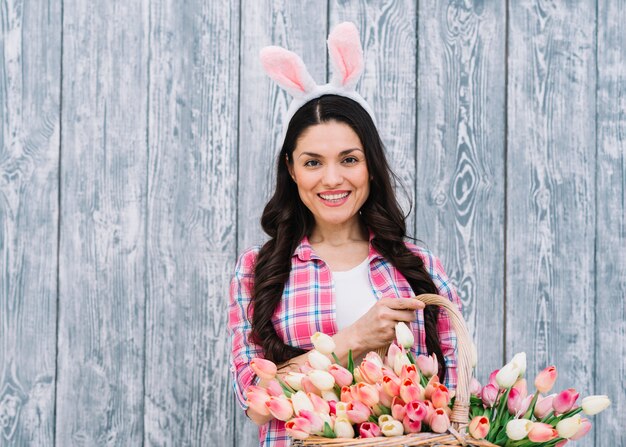 Retrato de una mujer sonriente con orejas de conejo en la cabeza con una canasta de tulipanes sobre fondo de madera