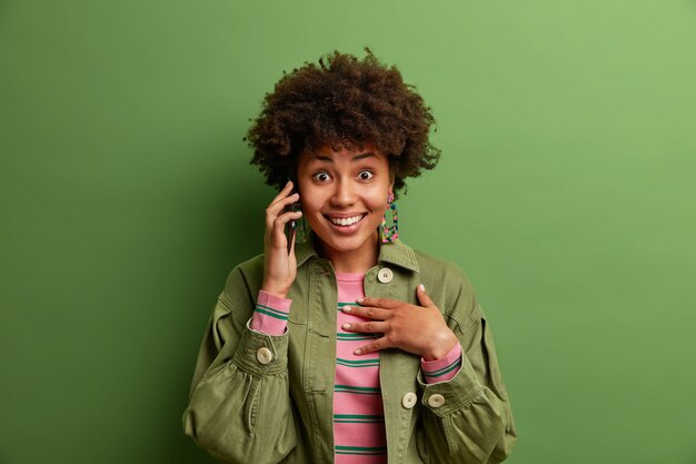 Retrato de mujer sonriente de pelo rizado habla a través de un teléfono móvil, disfruta de una agradable conversación, viste una chaqueta de moda, posa