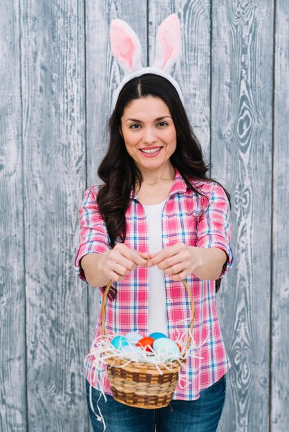 Foto gratuita retrato de la mujer sonriente que muestra la cesta de huevos de pascua contra fondo de madera