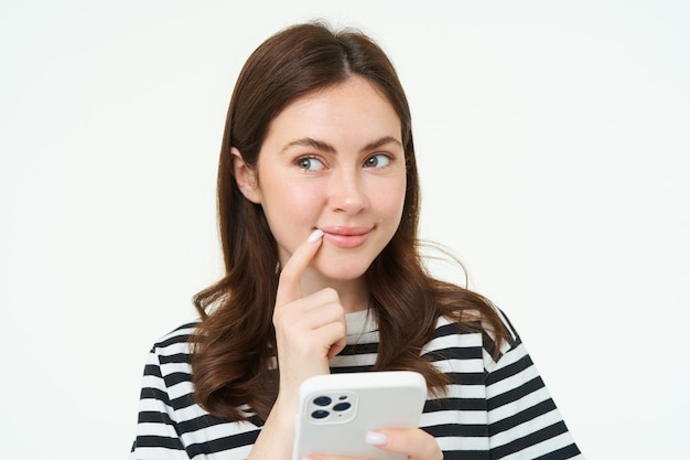 Foto gratuita retrato de una mujer sonriente que piensa pedir algo en línea en una aplicación de teléfono inteligente sosteniendo un teléfono móvil