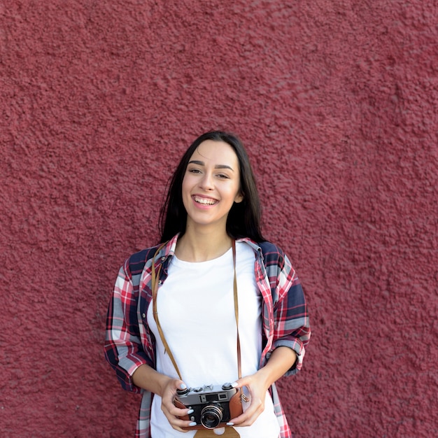 Retrato de la mujer sonriente que sostiene la cámara que se opone a la pared marrón