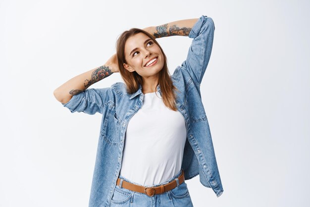 Retrato de una mujer sonriente y relajada cogida de la mano detrás de la cabeza mirando la esquina superior derecha descansando de ensueño teniendo tiempo libre de pie contra el fondo blanco